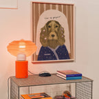 Orange lamp on a metal cabinet with framed art print of a cocker spaniel doing her hair with self-care quote on a striped background above it