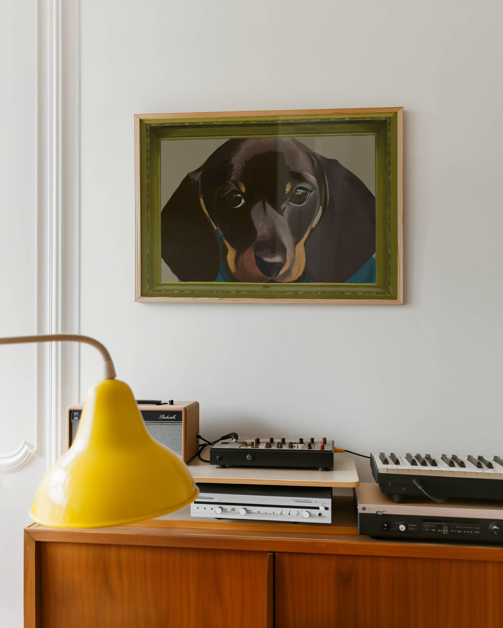 Framed portrait of a dachshund above vintage electronic equipment on a wooden cabinet